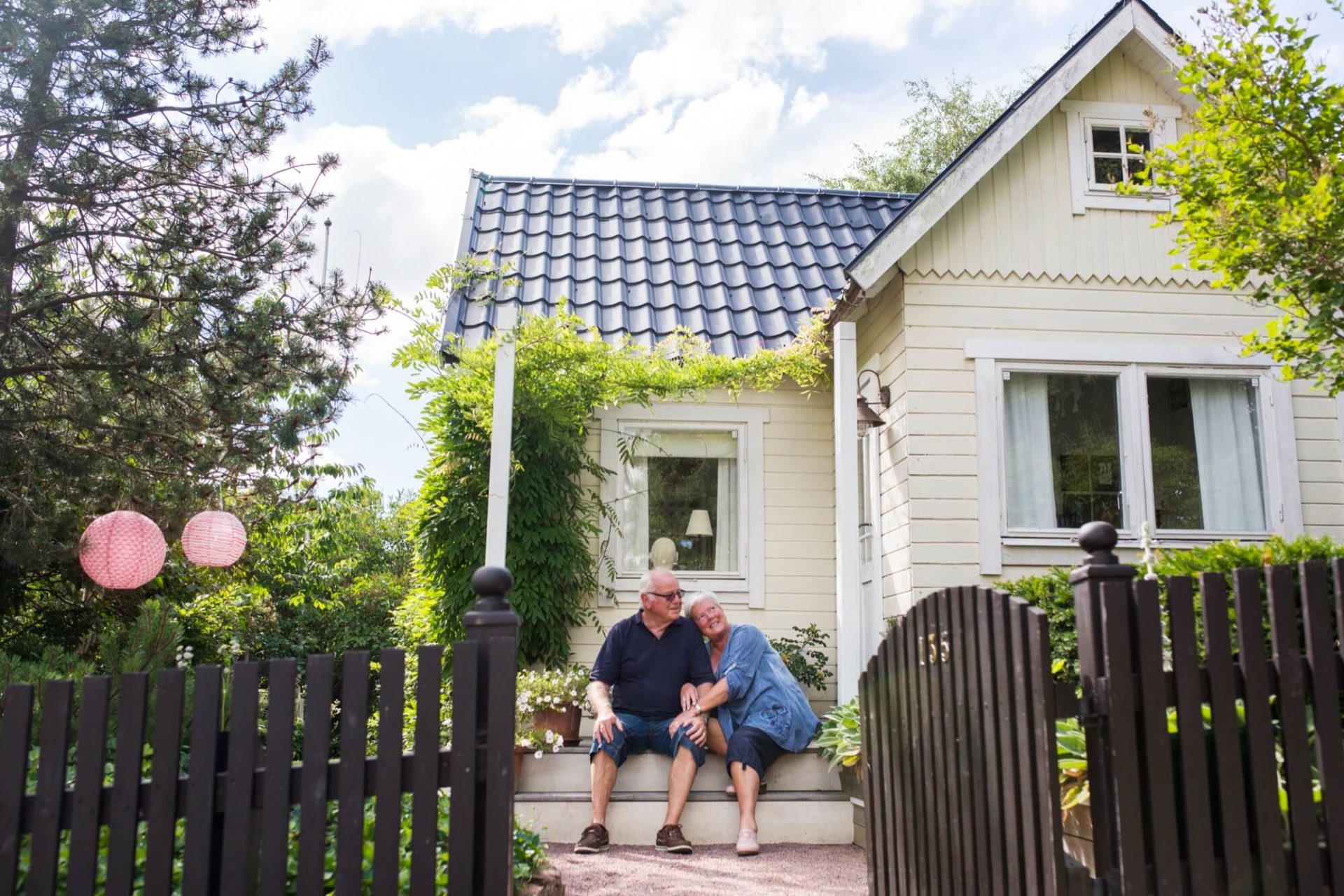 Ett äldre par sitter bekvämt på verandan till sitt charmiga trähus i ljusgul färg med ett svart tak, omringade av lummig grönska